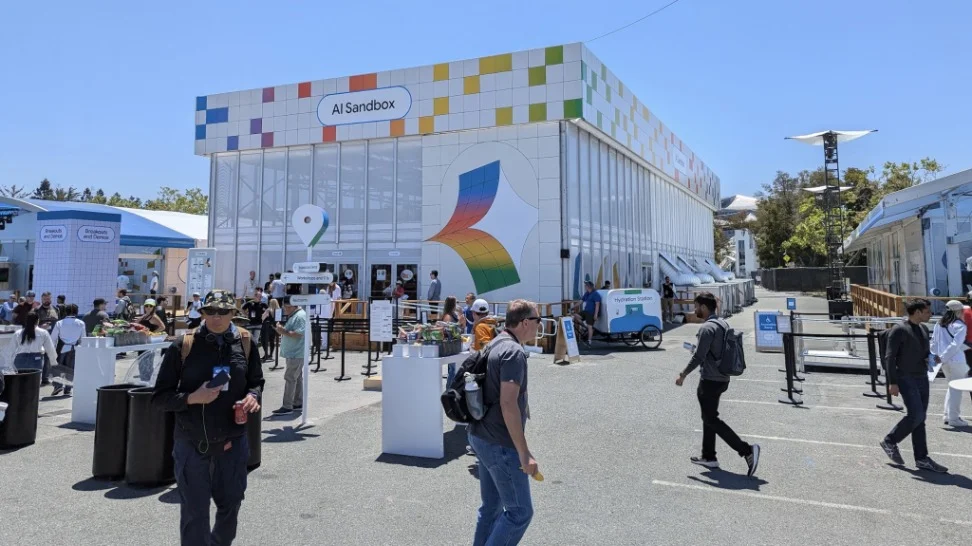 Photo of people walking around a parking lot, many wearing badges and backpacks. A giant, square building in the center has a banner that reads “AI Sandbox” at the top. The building is white with primary color squares dotting it.