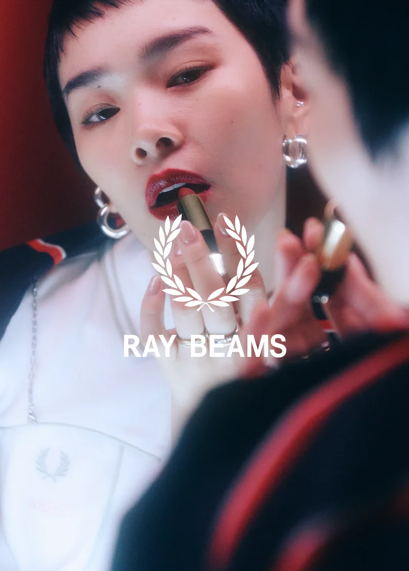 Fred Perry x RAY BEAMS: Japanese woman with short hair puts red lipstick on in front of a mirror, she wears silver hoop earrings and the white corset-style Ray BEAMS Fred Perry Shirt