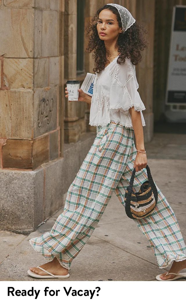 A woman wearing an embroidered white top, lace head scarf, plaid pants and sandals carrying a handbag.