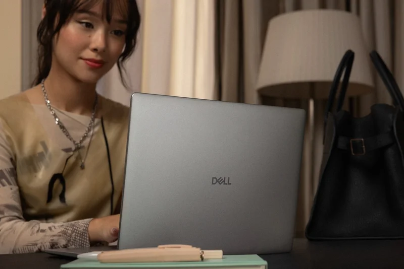 Stylish woman working at her home desk using a Dell 14 Plus laptop, with a notebook and handbag in a warm, modern setting.