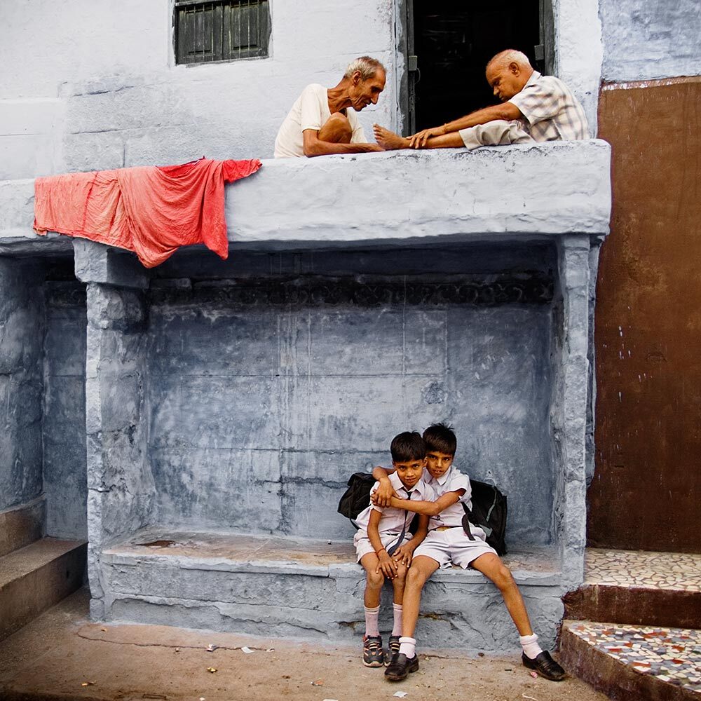 Two young boys embracing and looking at the camera while seated beneath a concrete balcony, upon which two elderly men are playing chess.
