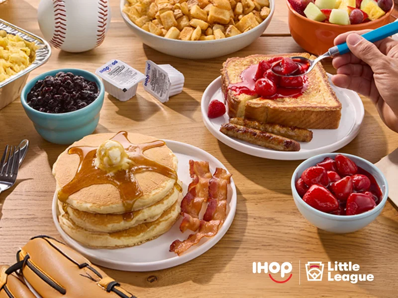 An assortment of IHOP breakfast items including pancakes, bacon and French Toast. A softball and mitt are on the table, along with the IHOP and Little League logos.