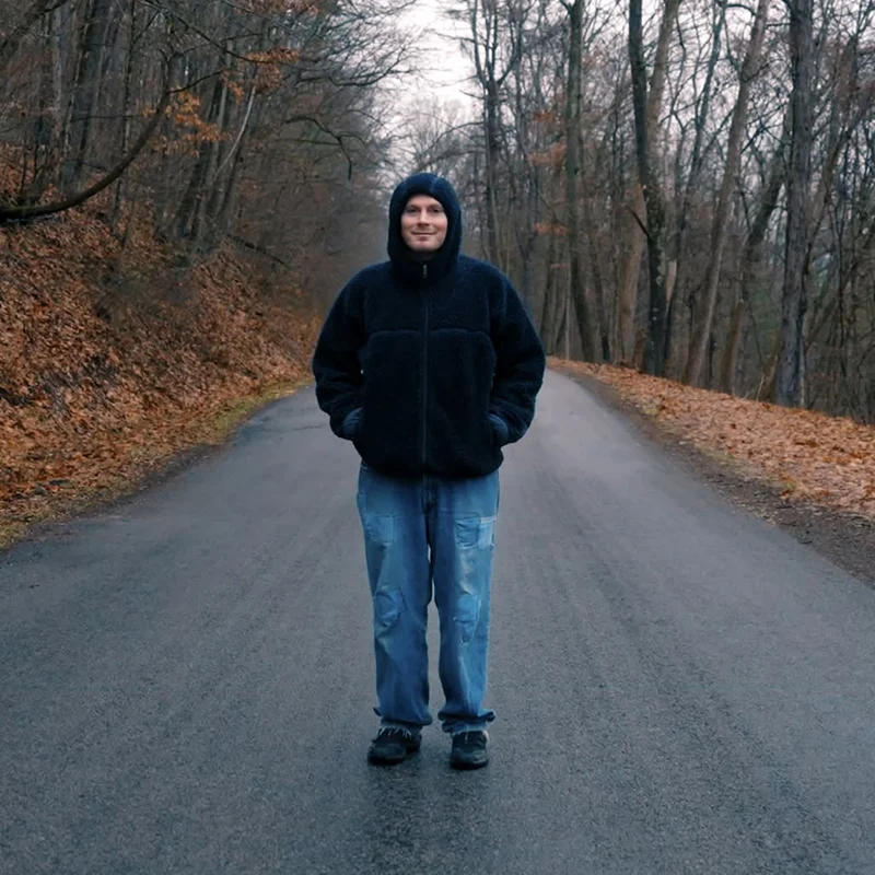 A person wearing jeans and a hoody stands in a road on a grey day surrounded by fallen leaves.