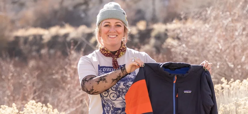 A person stands outside and holds up a repaired jacket with an orange patch on the upper arm.