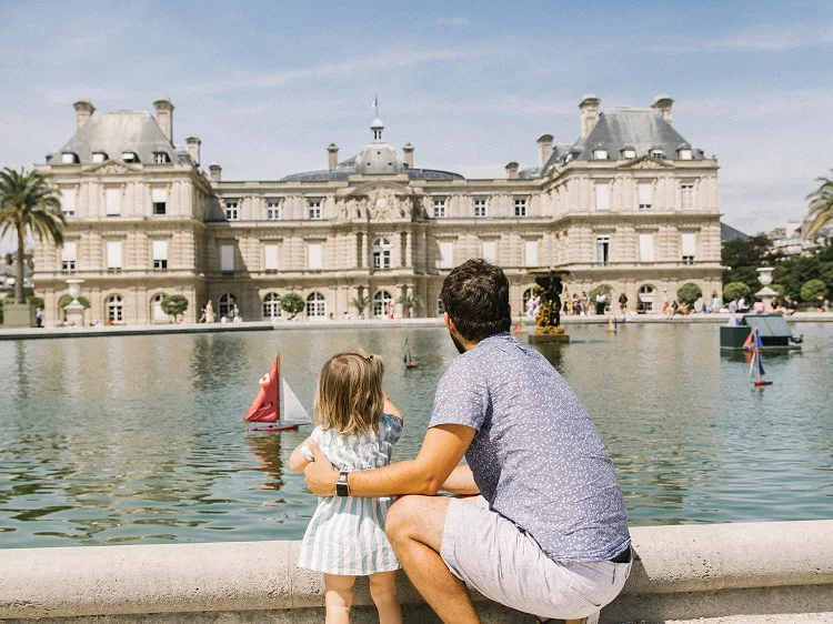 A father and young daughter stand by the edge of a large fountain pond, watching toy sailboats float in the water. Behind them is the historic Luxembourg Palace in Paris, France, under a bright blue sky.