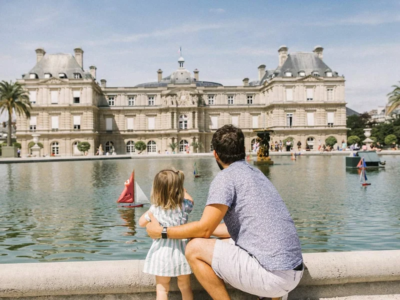 A father and young daughter stand by the edge of a large fountain pond, watching toy sailboats float in the water. Behind them is the historic Luxembourg Palace in Paris, France, under a bright blue sky.