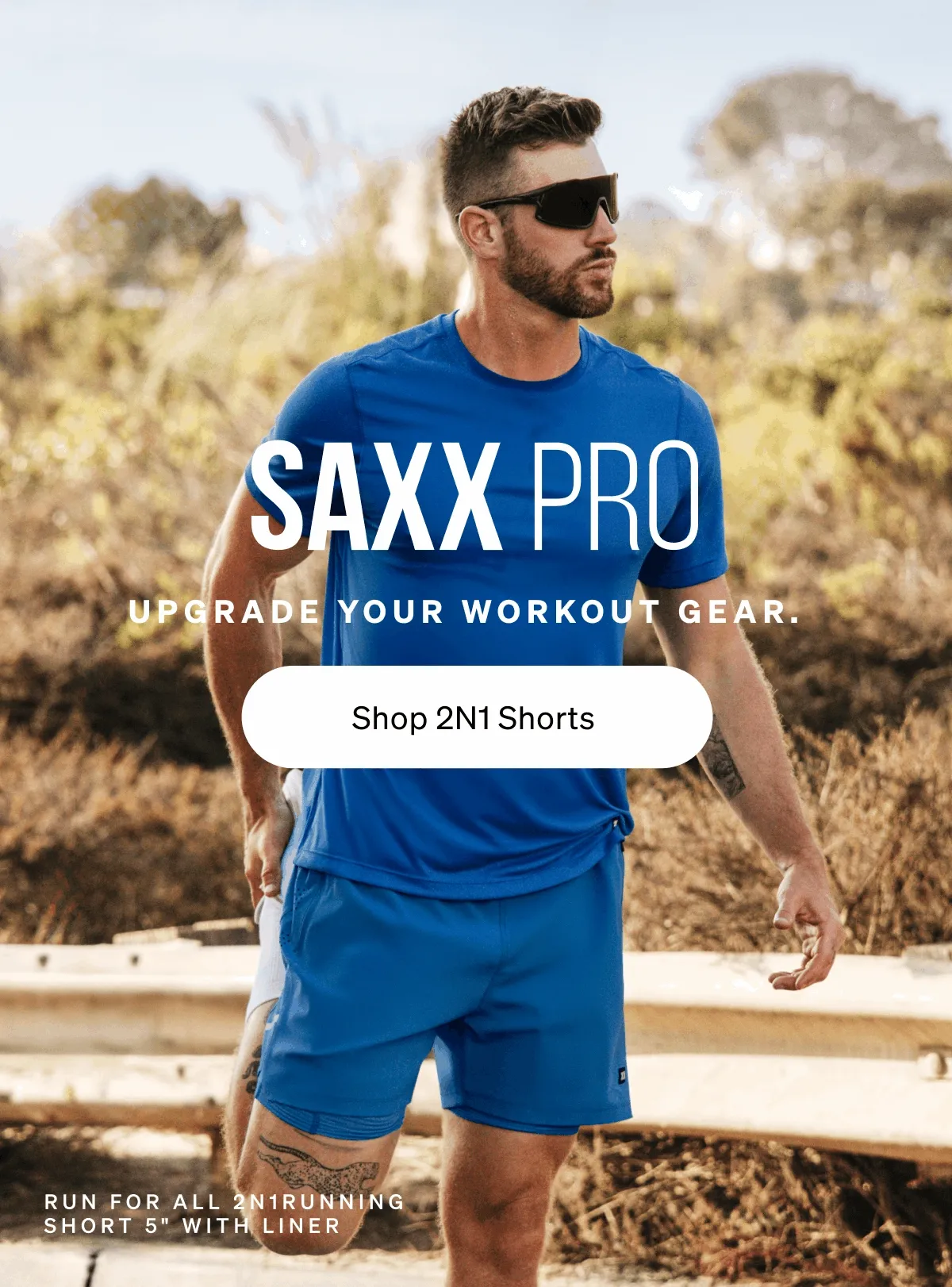 Man in blue athletic shirt and shorts stretching outdoors. Text reads “SAXX Pro. Upgrade your workout gear. Shop 2N1 Shorts.”