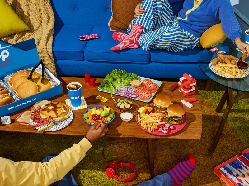 A casual scene with a person lounging on a blue couch, eating chicken tenders and fries on a table beside them. There is a large coffee table in the center of the room with IHOP catering on it. Another person sitting on the floor eats from a fruit bowl on the table.