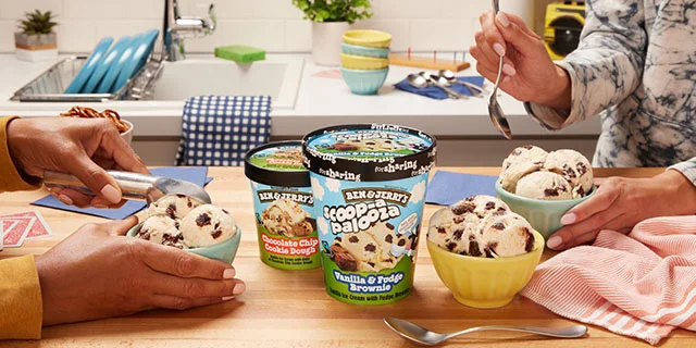 Kitchen setting, two people eating ice cream from bowls. On the table is a tub of Ben & Jerryt's Scoop-apalooza Vanilla & Fudge Brownie ice cream and a smaller pint of Ben & Jerry's Chocolate Chip Cookie Dough ice cream.