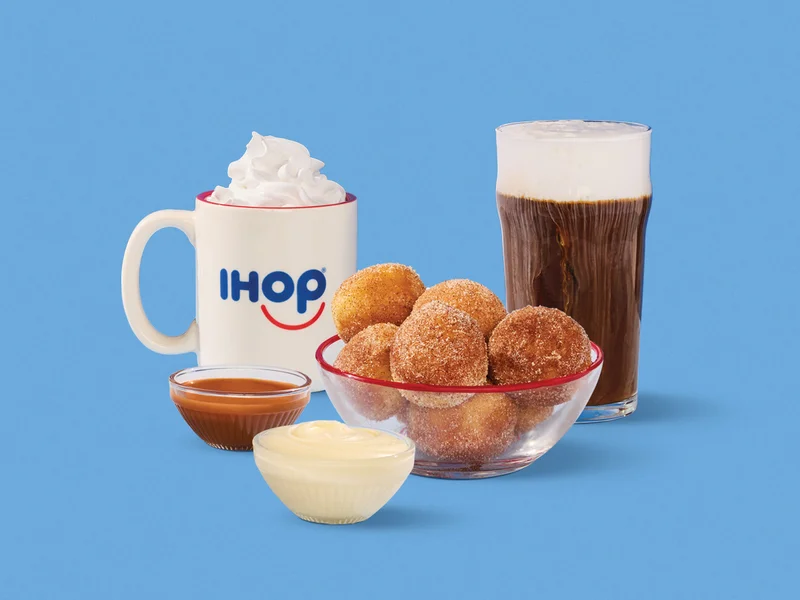 A mug of hot chocolate in an IHOP mug with whipped topping, alongside a cold brew coffee, Cinn-A-Dippers™, and two dipping sauces, set against a blue background