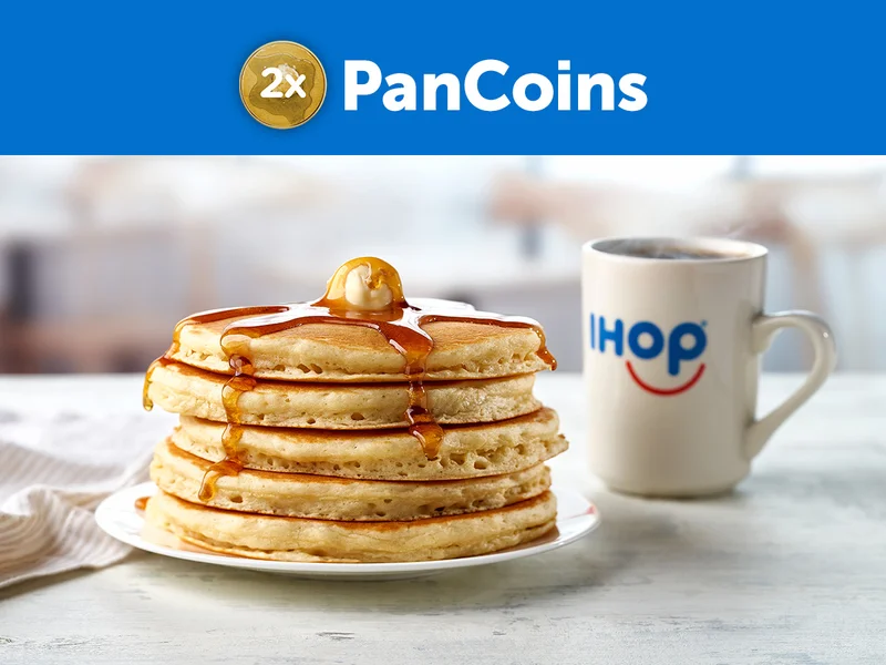 A full stack of buttermilk pancakes on a plate, topped with butter and syrup, beside an IHOP mug with coffee on a granite countertop