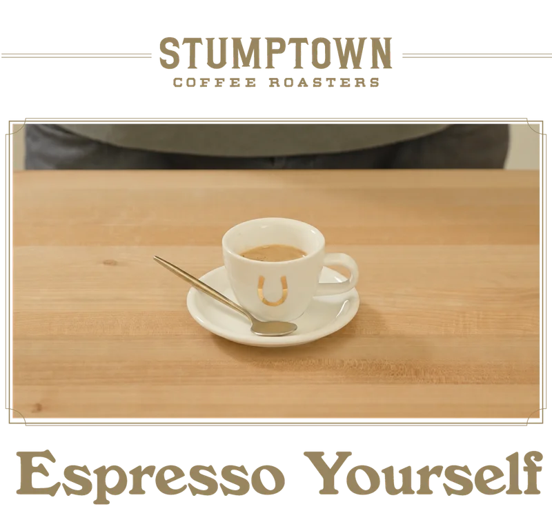 A small white espresso cup with a gold horseshoe logo sits on a saucer with a spoon on a light wood table. Text below reads “Espresso Yourself.”