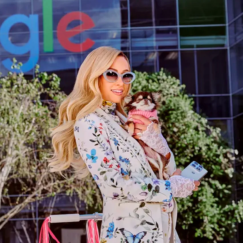 Paris Hilton holding a Chihuahua and smartphone in front of a Google building.
