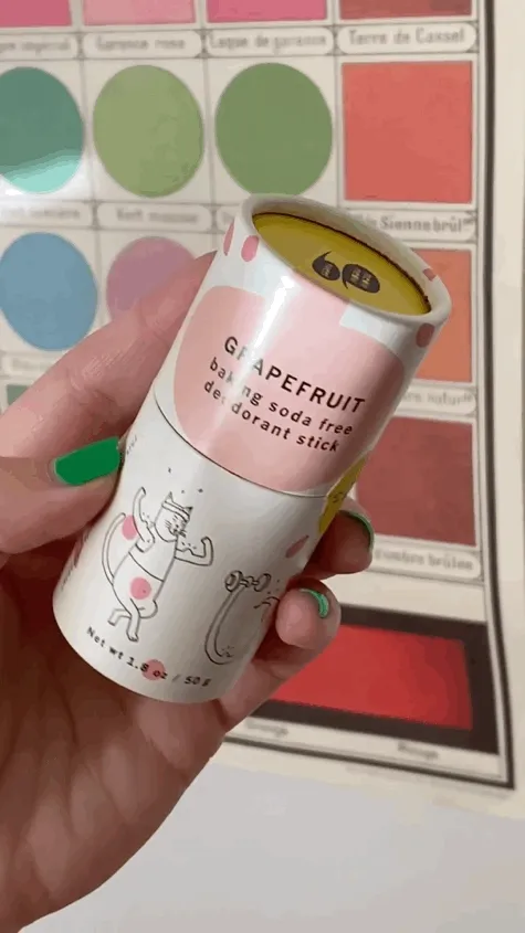 A light skinned person with brown hair, green nails and a red sweater opens a deodorant stick and applies it to their armpits