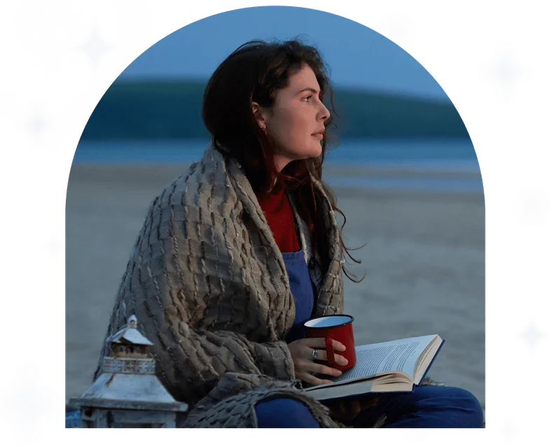 A woman sits on a beach at dusk beside a small camp fire and looks out to sea.