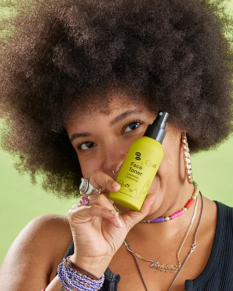 A Black person with an afro hides smiling behind a green bottle of toner