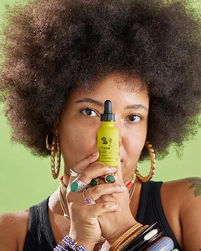 A Black person with an afro hides smiling behind a green bottle of oil