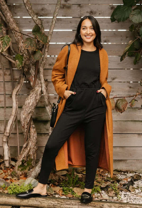 brunette woman wearing black romper and tan wool coat in front of wooden fence