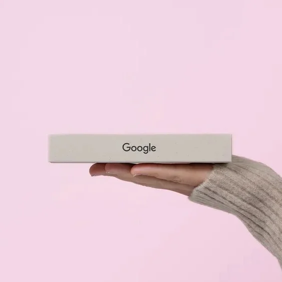 A hand balancing a small pink box that reads Google against a pink wall.