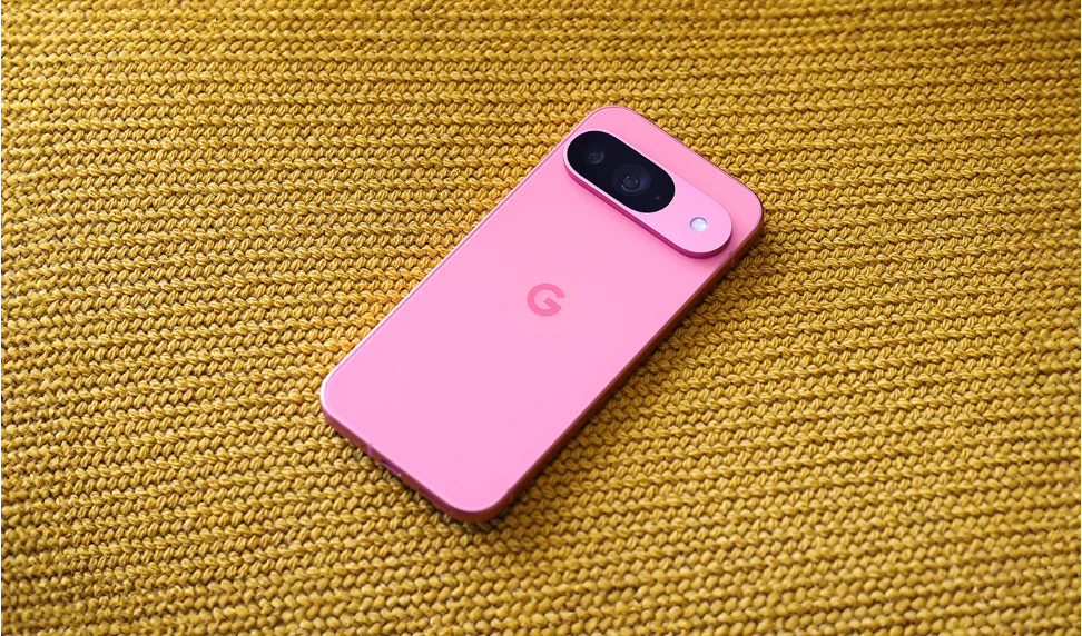 A pink Pixel 9 phone slightly tilted to the right, lying screen-down on a yellow knit fabric. The phone’s back is visible, showing its pill-shaped Camera Bar at the top of the device and a slightly darker shaded pink “G” in the center of the phone.