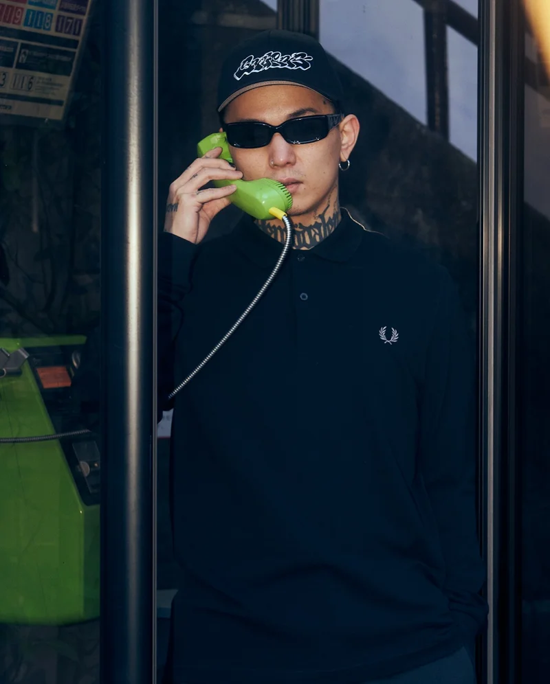 Asian male model wearing a black long sleeved fred perry shirt in a phone booth on the phone