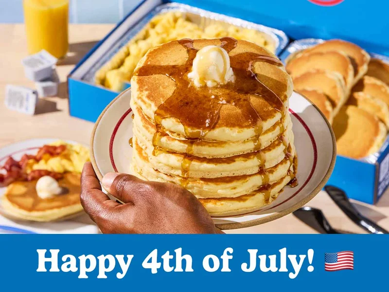 A stack of pancakes on a plate is being held above a catering tray of pancakes and scrambled eggs on a table below it