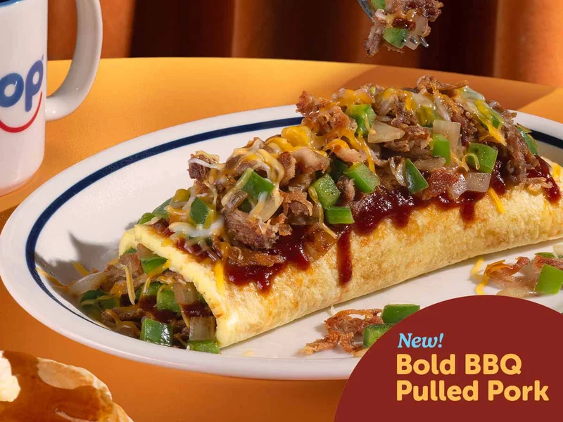 The Bold BBQ Pulled Pork Omelette is on an IHOP plate, on a table in a booth, with a mug of IHOP coffee and some pancakes cropped out of frame.