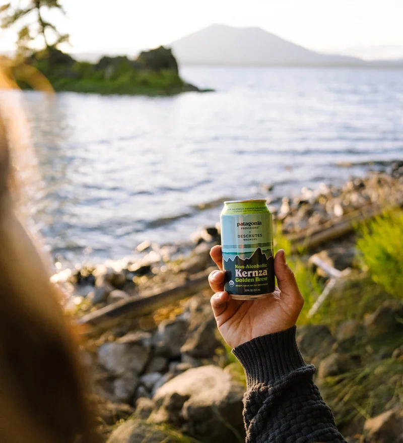 A hand holds up a can of non-alcoholic beer on the shore of a large body of water.