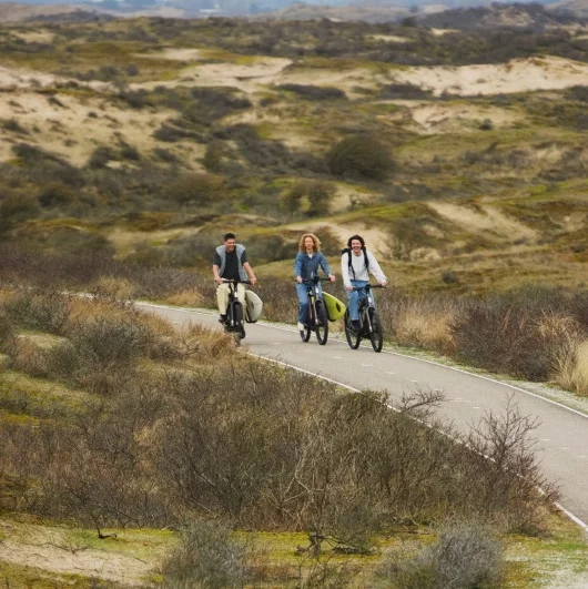 Three riders cycling together on Cowboy bikes through a winding path surrounded by open sandy hills and scattered shrubbery.