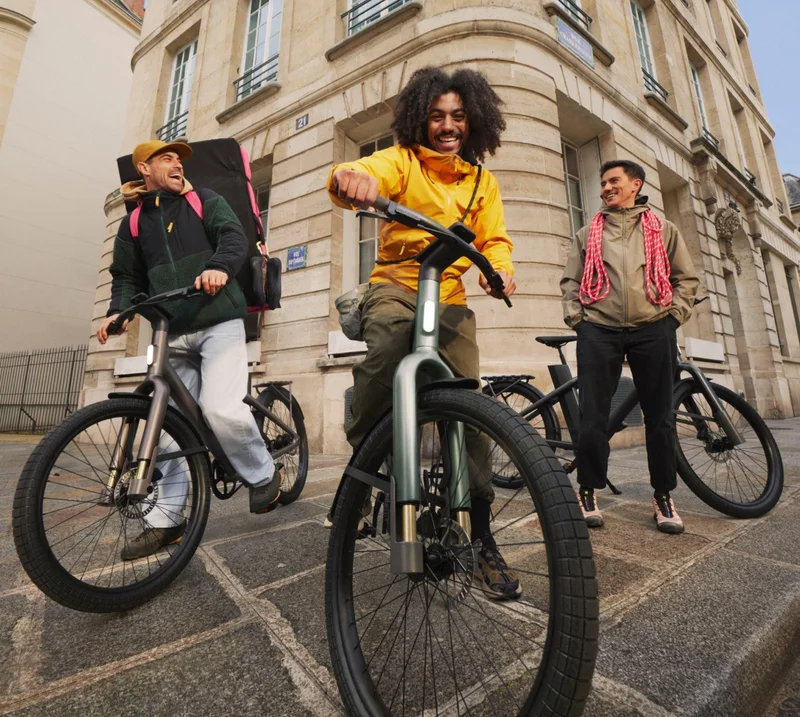 A group of three friends standing with their Cowboy bikes in a city setting. The person in the bright yellow jacket sits on a green bike, smiling, while the others lean casually against their bikes and chat.
