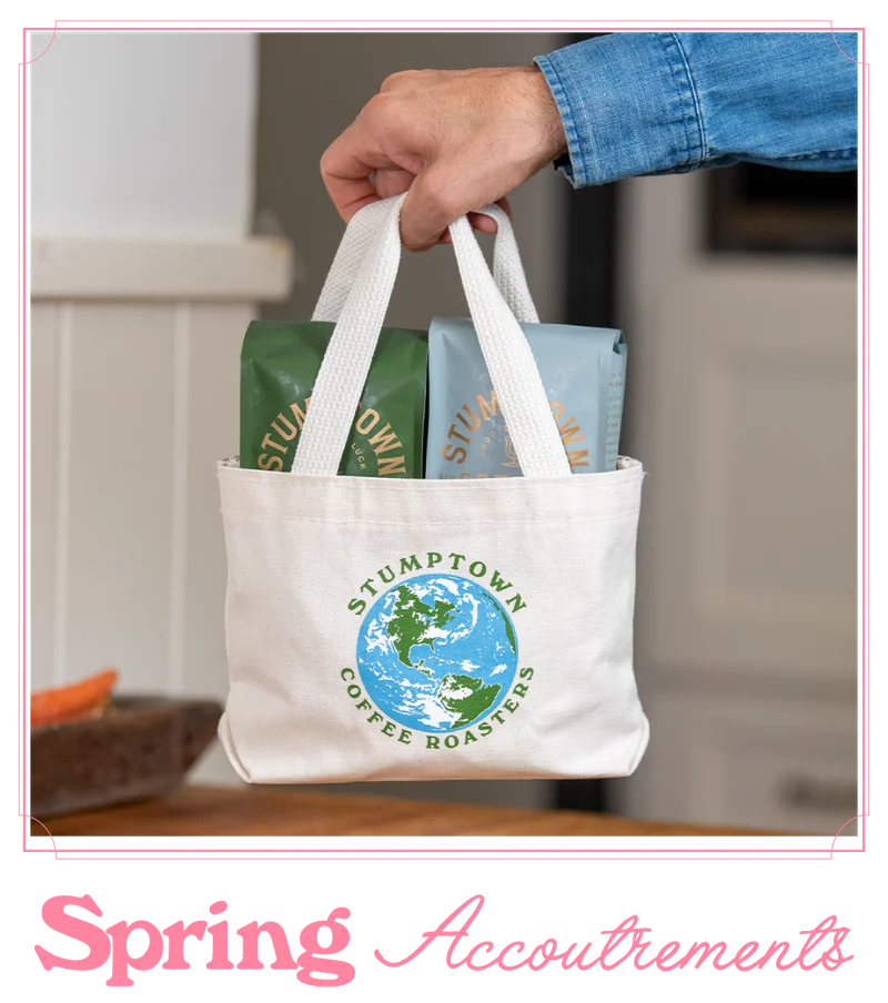 Hand holding a Stumptown Coffee Roasters mini canvas tote with a globe logo, filled with two coffee bags, with the headline “Spring Accoutrements” below.