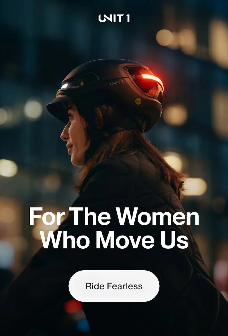 Woman riding at night wearing a UNIT 1 smart helmet with rear light illuminated and the headline “For The Women Who Move Us”