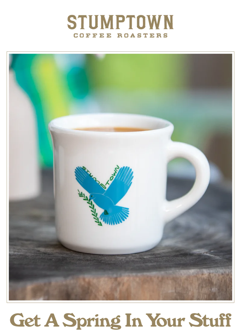 White Stumptown espresso mug with a blue dove illustration sitting on a wooden table. Headline reads: “Get A Spring In Your Stuff.”