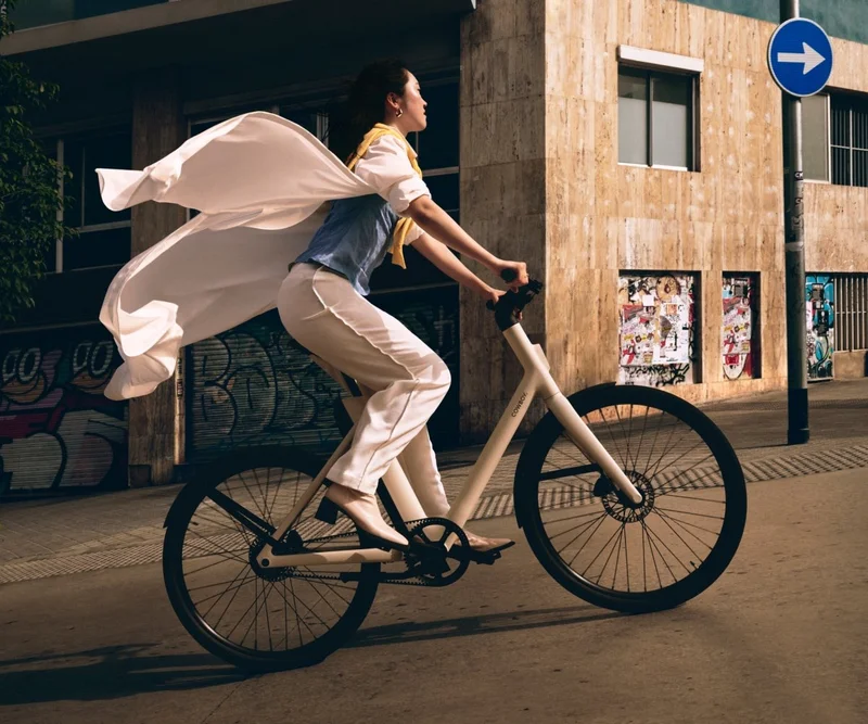 A photograph. A woman riding a Cowboy e-bike through an urban area, with her coat flowing dramatically behind her.