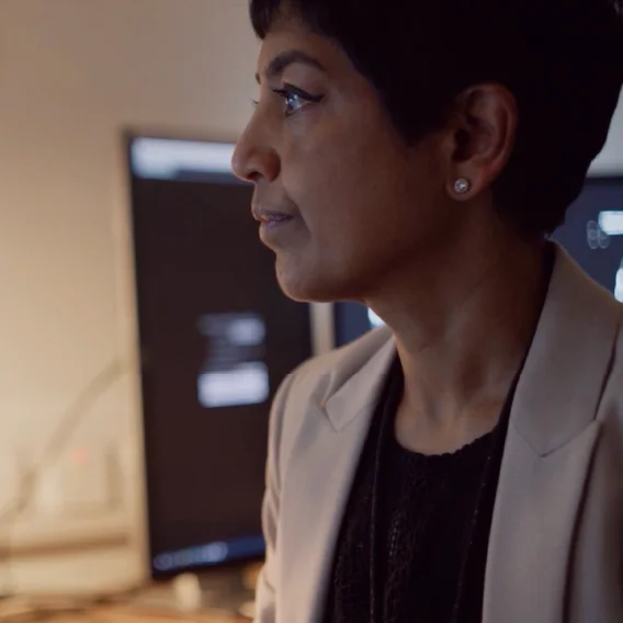 Profile of a woman with short dark hair and a light blazer looking at a computer monitor in a dimly lit office.
