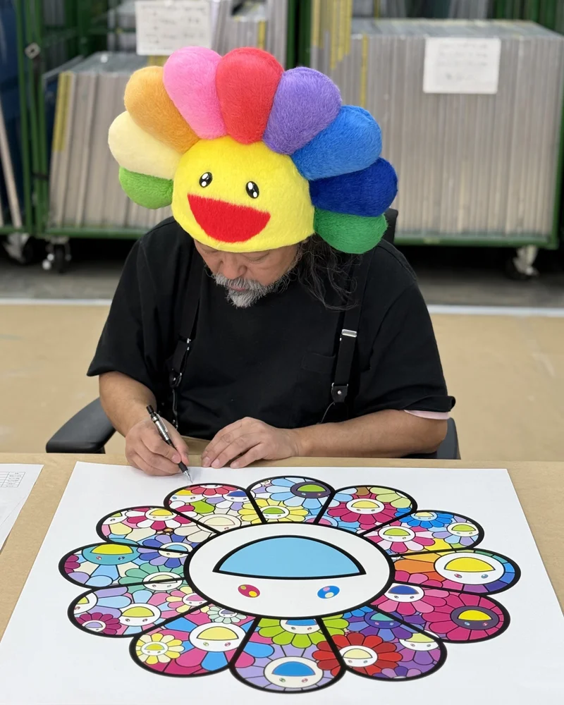 Artist Takashi Murakami dons a bright flower hat while signing a print