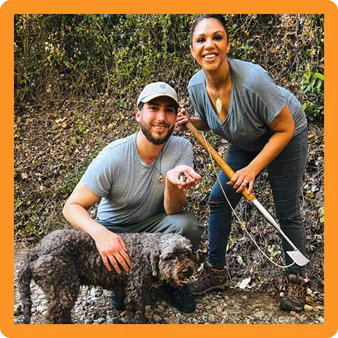 Image description: Two travelers smiling and posing with truffles, a dog, and a truffle-hunting tool.