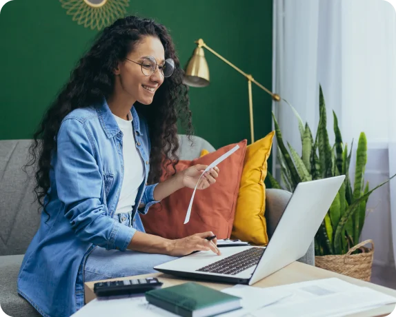 Person with long hair and glasses holding a receipt and working on a laptop in a living room