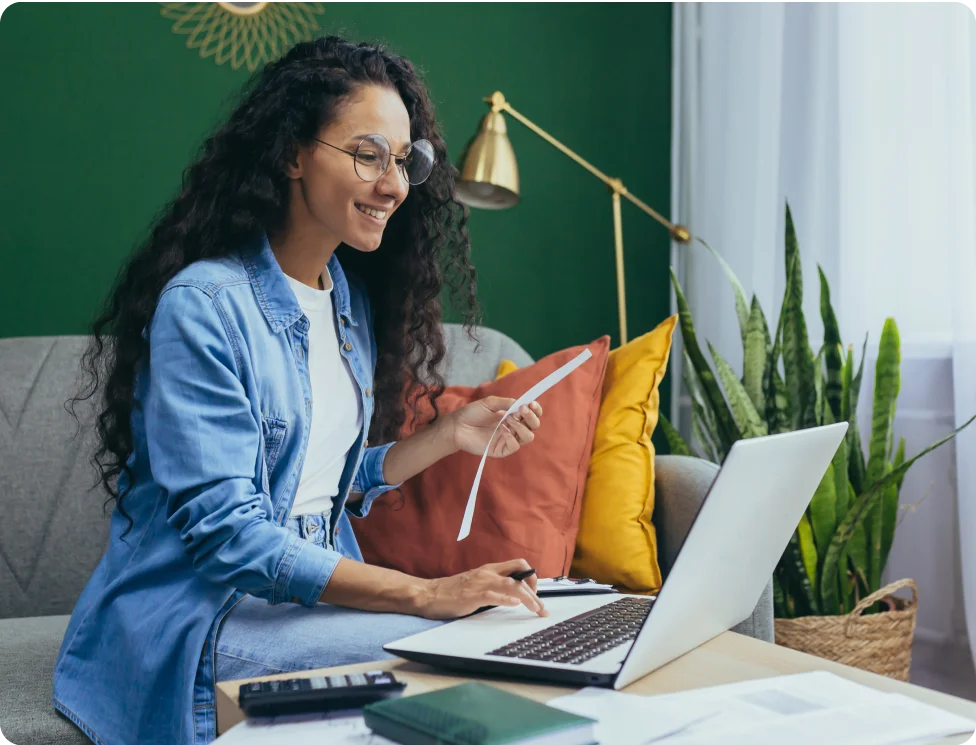 Person with long hair and glasses holding a receipt and working on a laptop in a living room