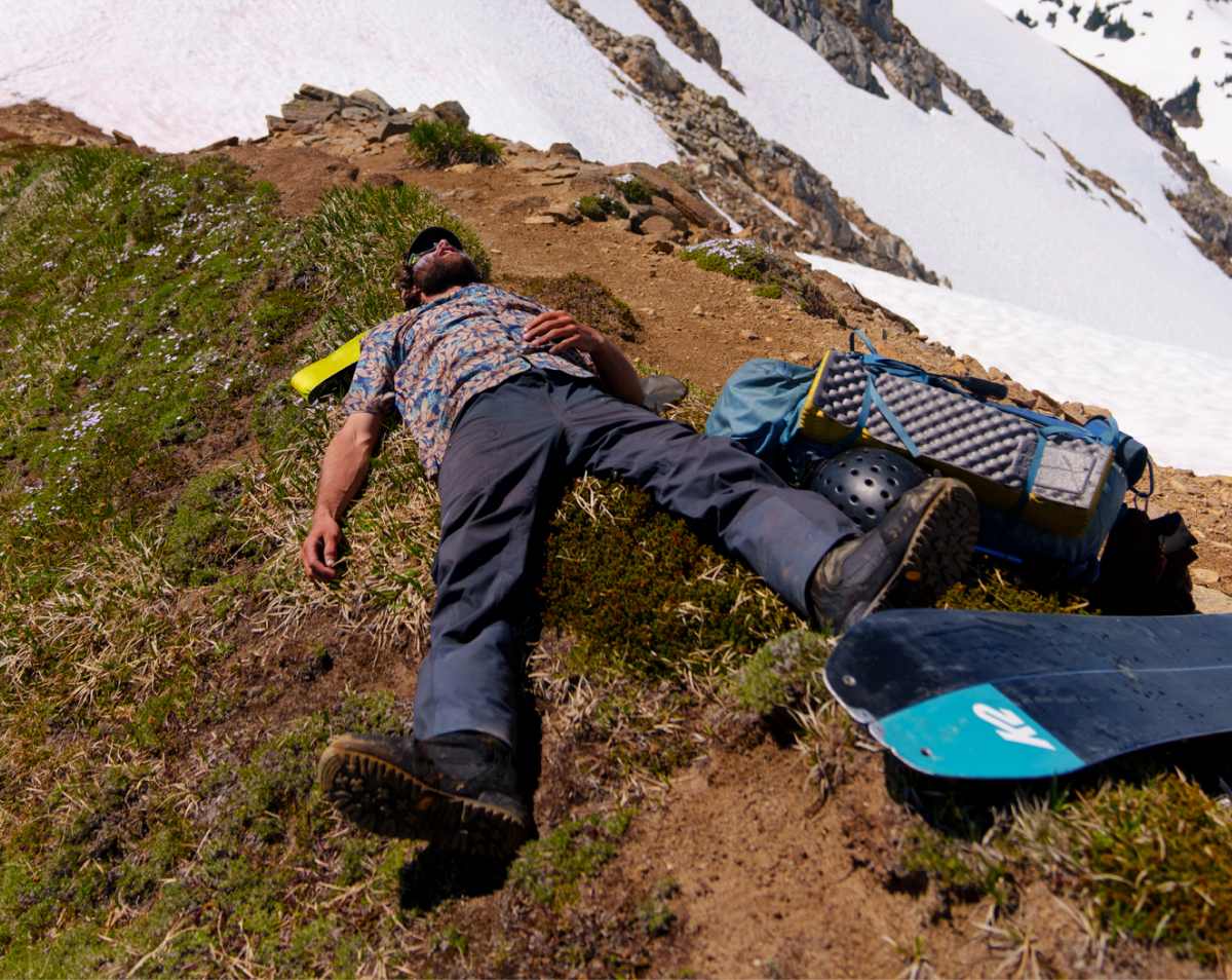 A person laying on the ridge of a mountain barely covered in snow with snowboard gear next to them.