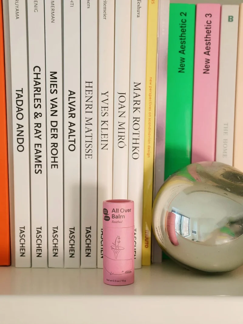 A pink paper tube of All Over Balm sitting on a bookshelf