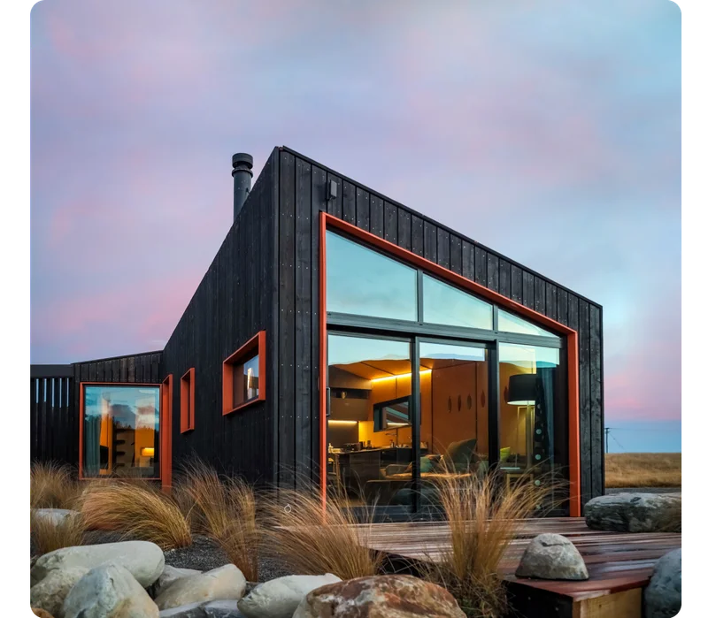 An elegant wood home with floor-to-ceiling windows nestled in the New Zealand plains. Dark, wood exteriors contrast the interior warmth of the home with soft lights and modern furniture.