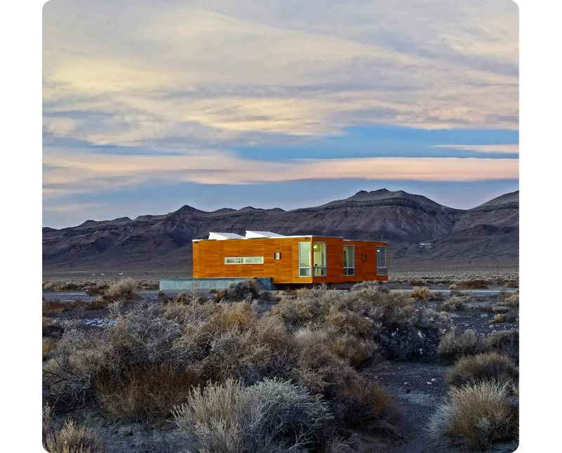 A modern home crafted from light wood sits in a remote field of shrubs with rolling brown mountains in the background.