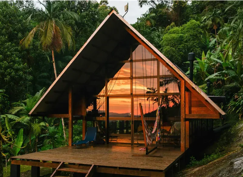 An elegant wood home with floor-to-ceiling windows is surrounded by lush greenery and palm trees. An in-round fire pit sits in front of the home, as well as a small deck with a blue chair and hammock.