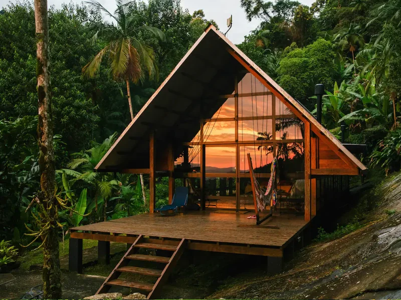 An elegant wood home with floor-to-ceiling windows is surrounded by lush greenery and palm trees. An in-round fire pit sits in front of the home, as well as a small deck with a blue chair and hammock.
