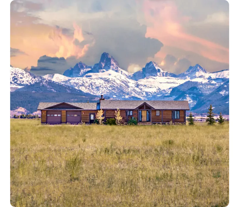 A meadow leads up to a brown, wood house situated directly in front of the Grand Tetons capped with snow.