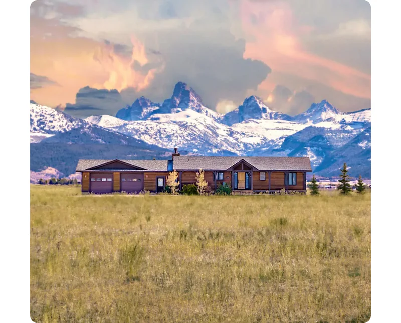 A meadow leads up to a brown, wood house situated directly in front of the Grand Tetons capped with snow.