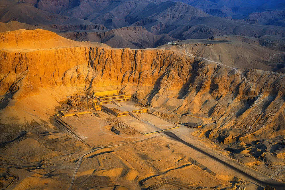 An aerial view of the Deer el Bahri temple site, in the desert, with a sun setting casting orange light on the sand.