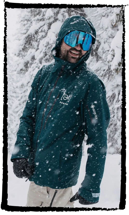 Snowboarder wearing the Men's [ak]® Cyclic Jacket in deep emerald in the snowy mountains