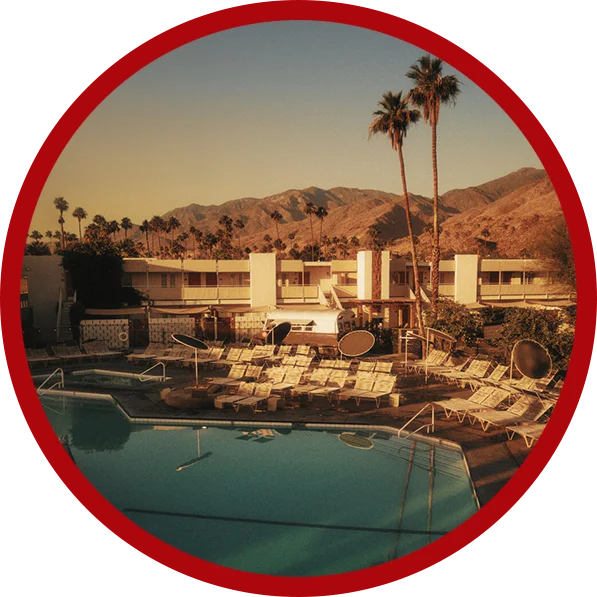 Scenic view of Ace Hotel & Swim Club Palm Springs: a serene pool area flanked by sun loungers, framed by palm trees and low-rise buildings, with the picturesque mountains of Palm Springs in the background.